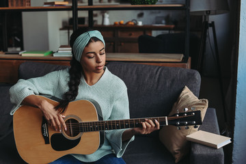 beautiful mixed race girl in turquoise sweater and headband playing acoustic guitar on sofa at home - Powered by Adobe