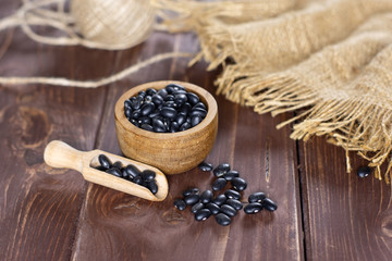 Lot of whole raw black turtle beans jute cloth and a scoop with wooden bowl on brown wood