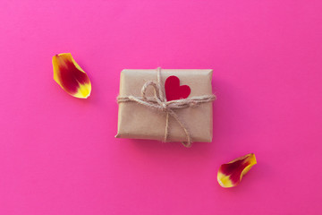 Gift in craft paper with red heart and red-yellow petals on the pink background. Trendy bright colors, minimal fashion stillife. Top view, flat lay, copy space