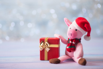 Closeup cute toy pig in Santa hat and scarf sitting on wooden table and holding festive gift box on light bokeh background