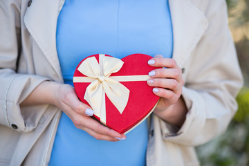 Young female hands holding gift box in shape of red heart with bow.