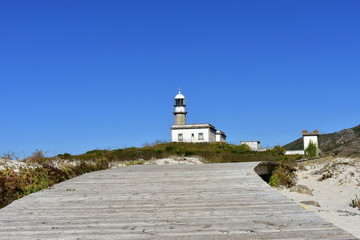 Beach with boardwalk and old abandoned lighthouse on a hill. Sunny day, blue sky. Galicia, Spain.