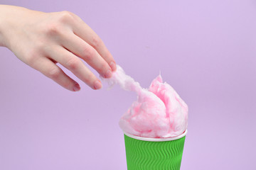 Bright pink candy cotton in plastick cup in woman hand on lilac background.