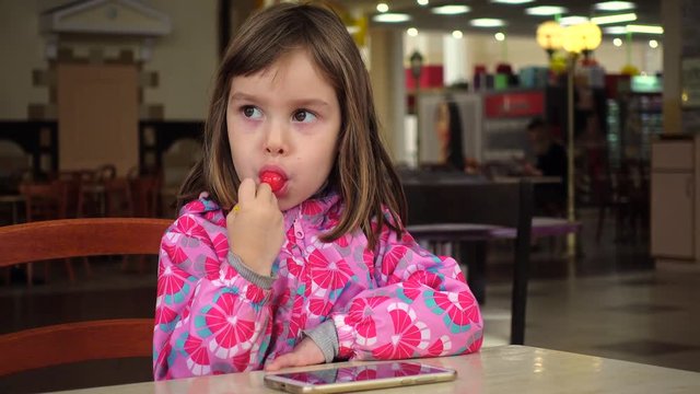 A little girl, a child of 4-5 years old, eats chupa chups, sitting at a table on a food court.