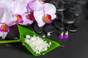 spa composition of lilac orchid with drops, aroma incense cones and sea salt on leaf over black zen stones