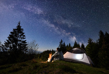 Man tourist sitting alone near illuminated tent at burning campfire on grassy valley, enjoying night blue starry sky with Milky way, pine trees forest on background. Beauty of nature, tourism concept