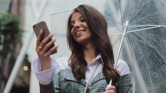 Beautiful young business woman using smartphone on the street in rainy weather, smiling, holding umbrella, communication concept