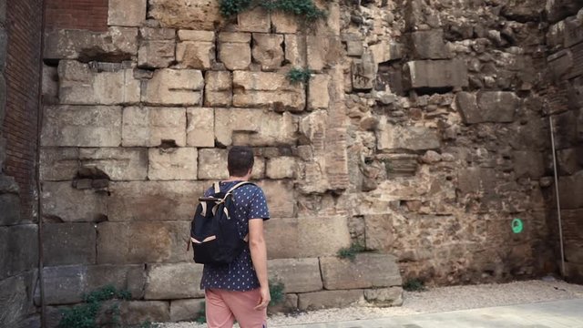 Male traveler is viewing ancient stone walls in center of Barcelona in summer day. He is looking up and approaching to castle ruins, admiring by them