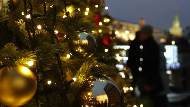 Holidays in Moscow. In the square a Christmas tree glows, decorated with balls and beautiful Christmas toys close-up. Kremlin tower, pedestrians and travellers walk on blurred background