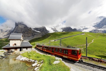 A cog wheel train travels on Jungfrau Railway from Jungfraujoch station (top of Europe) to Kleine Scheidegg on a green hillside with a snow shed tunnel on a cloudy day in Bernese Oberland, Switzerland