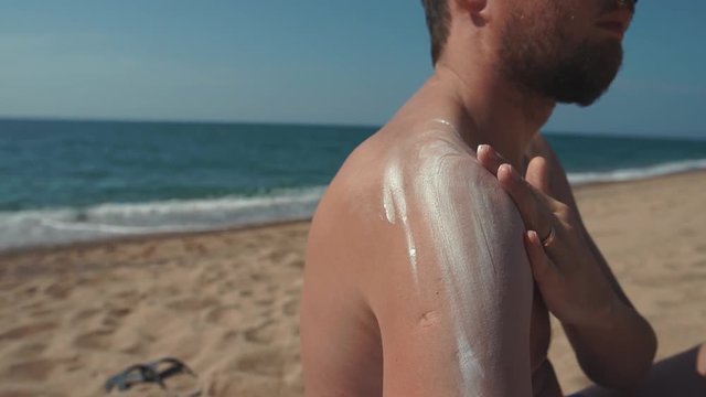 Close-up shot of a man applying sunblock lotion on his shoulders and rubbing in. Man protects his skin on the beach.