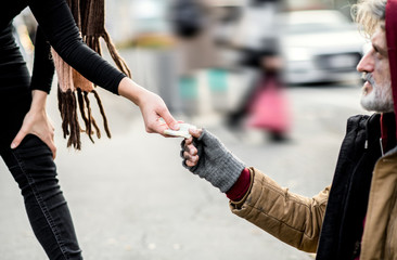 A midsection of woman giving money to homeless beggar man sitting in city.