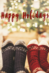 Happy Holidays text on stylish festive socks on couple legs at golden christmas tree  lights in festive room. Season's greetings card. Merry Christmas and Happy New Year