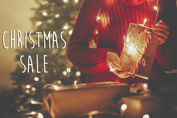 Christmas big sale text. Holiday discount offer. Hands holding christmas present in lights in evening festive room under tree in illumination. Shopping and sales