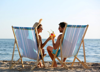 Obraz premium Young couple with cocktails in beach chairs at seacoast