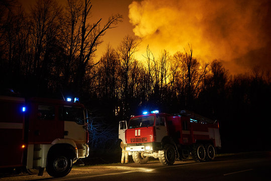 Forest Fire With Smoke And A Road With Fire Engines At Night, View From The Distance