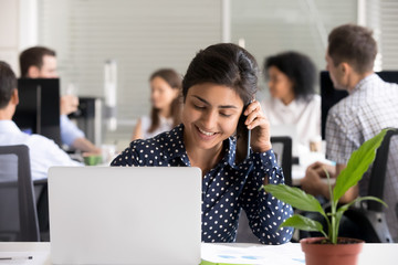 Smiling beautiful Indian female employee talking on phone at workplace, consulting, young businesswoman holding smartphone in hands, making call, having pleasant conversation, chatting by mobile phone