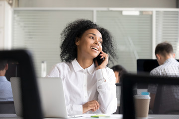 Smiling African American woman making call, talking on phone with client or customer at workplace,...