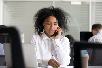 Focused African American woman making notes on sticker, clarifies information, talking on phone,...