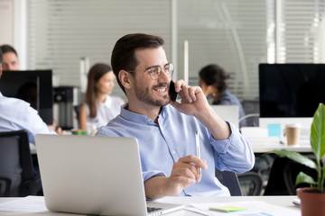 Smiling friendly man talking on phone in office, having pleasant conversation with client or customer, receiving good news or offer, marketing manager making business call, chatting with friend