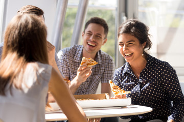 Indian excited woman laughing at funny joke, eating pizza with diverse colleagues in office, happy multi-ethnic employees having fun together during lunch, enjoying good conversation, emotions