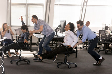 Excited diverse employees having fun together in office, riding on chairs at work, enjoying break,...