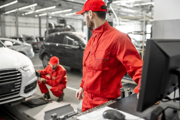 Two handsome auto mechanics in red uniform making wheel alignment with professional tools and...