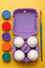 Easter composition. Set of four white eggs in a purple box, multi-colored jars of paint and a brush for drawing on a yellow background
