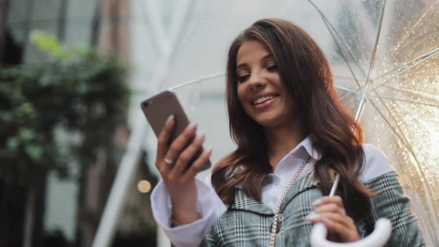 Beautiful young business woman using smartphone on the street in rainy weather, smiling, holding umbrella, communication concept