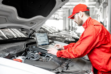 Handsome auto mechanic in red uniform doing engine diagnostics with computer in the car service