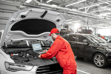 Handsome auto mechanic in red uniform doing engine diagnostics with computer in the car service