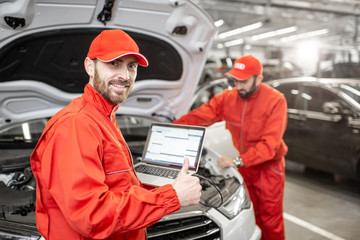 Two auto mechanics in red uniform doing engine diagnostics with computer in the car service