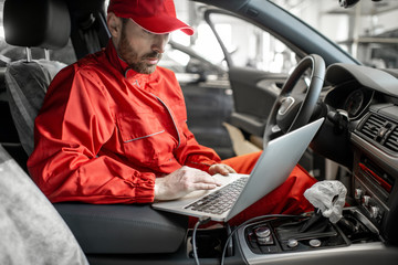Auto mechanic in red uniform diagnosing car with computer sitting on the driver seat at the car...