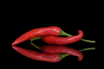 Two bright red chilli peppers with green stalks on a black background with a clear and rich reflection in the glass