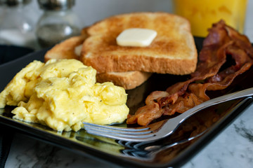 Scrambled eggs with bacon and toast with fork on black plate.  Salt, pepper, and orange juice in the background.