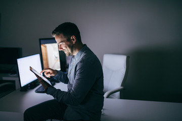 Smiling Caucasian bearded businessman using tablet while sitting on the desk in the office late at night.