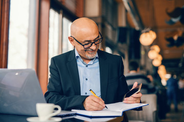 Bearded senior businessman looking at smart phone and writing tasks in agenda while sitting in...