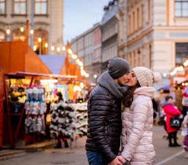 A young romantic couple wearing winter clothes hold hands and kissing while standing in evening street with Christmas fair