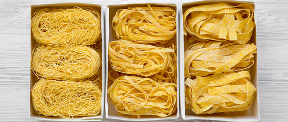 Set of various uncooked pasta in paper boxes on a white wooden surface, overhead view. From above, top view. Closeup.