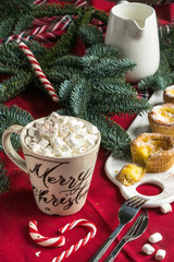 White big mug with cocoa and marshmallows on a background of red napkin, sweets and spruce branches. New Years concept, comfort and warmth.