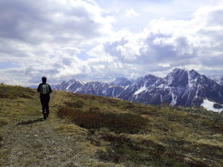 Fototapeta na wymiar persona che cammina su sentiero di alta montagna delle alpi con sfondo dolomiti innevate e nuvole in cielo