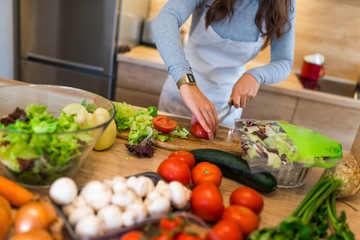 Delightful and happy young woman stands and cooks in kitchen. She cuts lettuce on wood board. Girl has many ingrediends on table.