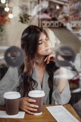 Gorgeous young woman with cup of coffee sitting in cafe and relaxing at window in city street. Stylish hipster girl with beautiful hair enjoying time with coffee