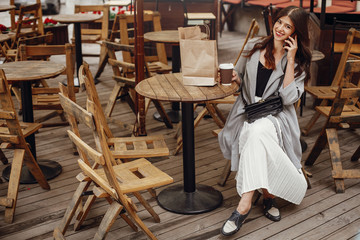 Fototapeta na wymiar Gorgeous young woman talking on phone and smiling, with cup of coffee and paper bag on wooden rustic table on terrace in city street. Stylish hipster girl with beautiful hair and perfect smile