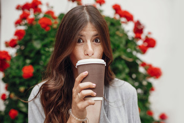 Stylish hipster girl with beautiful hair holding coffee cup and smiling in city street on background of flowers. Gorgeous happy young woman with funny emotion enjoying time .