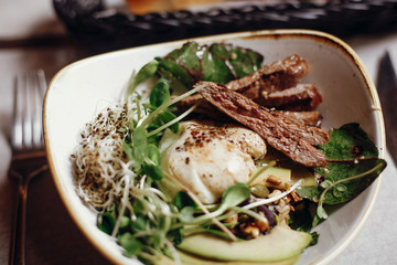 Delicious salad with juicy grill steak, avocado, sprouted greens, leaves, egg pouch, nuts in stylish bowl on table at cafe in city street. Tasty modern salad with duck meat and vegetables