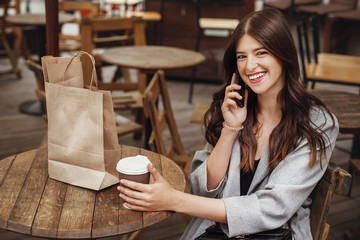 Stylish hipster girl with beautiful hair and smile talking on phone, with coffee cup and bag on wooden table on terrace in city street. Gorgeous happy young woman enjoying time and smiling