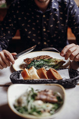 Hands holding fork and knife and eating delicious juicy steak with grilled cabbage,tomatoes and cheese on table at cafe in city street. Man tasting bbq with vegetables in restaurant