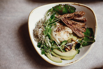 Delicious salad with juicy grill steak, avocado, sprouted greens, leaves, egg pouch, nuts in stylish bowl on table at cafe in city street. Tasty modern salad with duck meat and vegetables