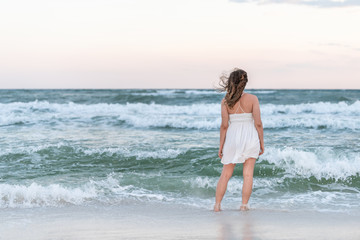 Fototapeta na wymiar Young woman back standing in white dress on beach sunset in Florida panhandle with wind, ocean waves, tan skin
